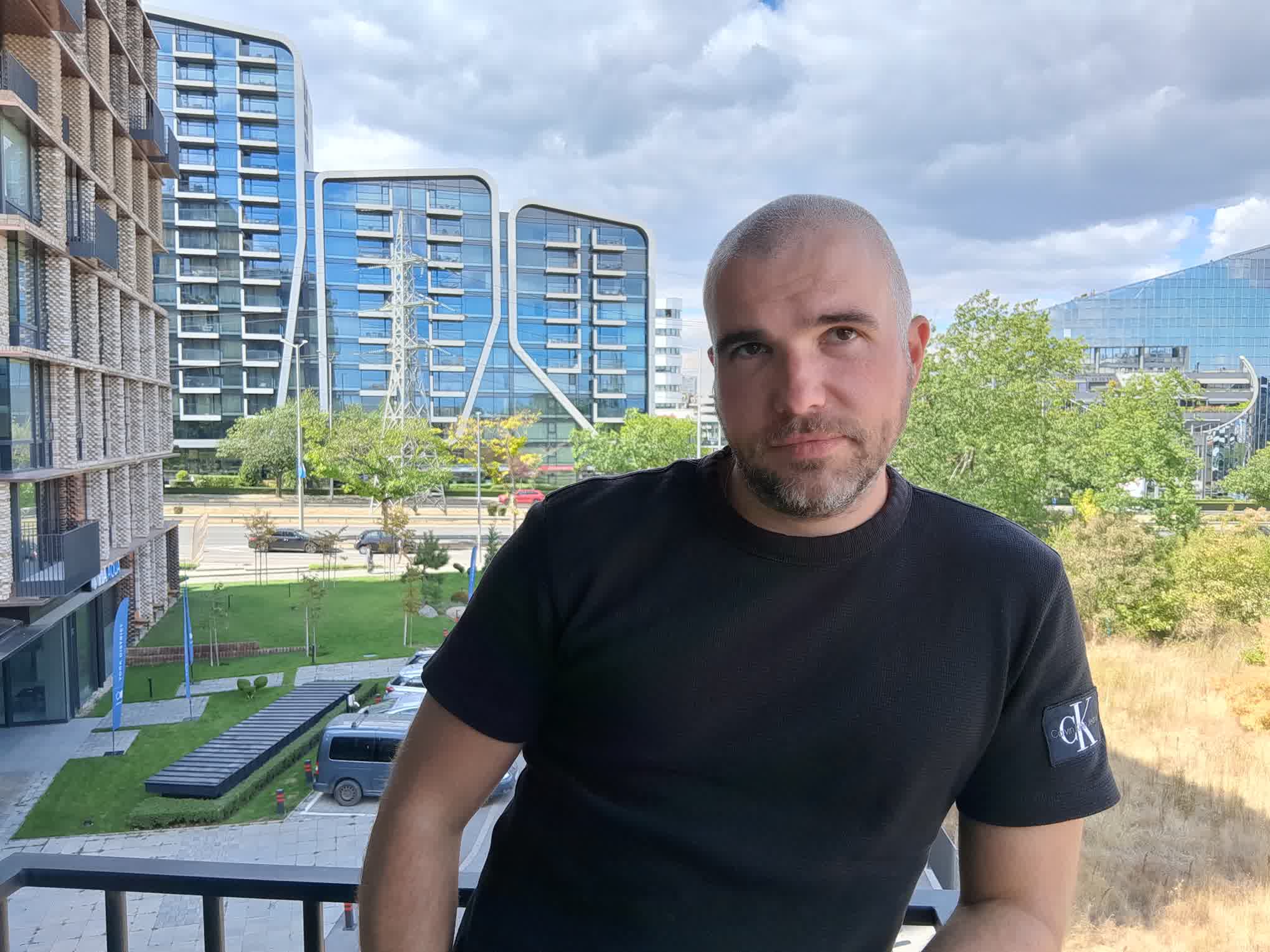 gsmarena_4101 Man in a black t-shirt taking a selfie on a balcony, with blue glass apartment/office buildings behind him under a cloudy sky.