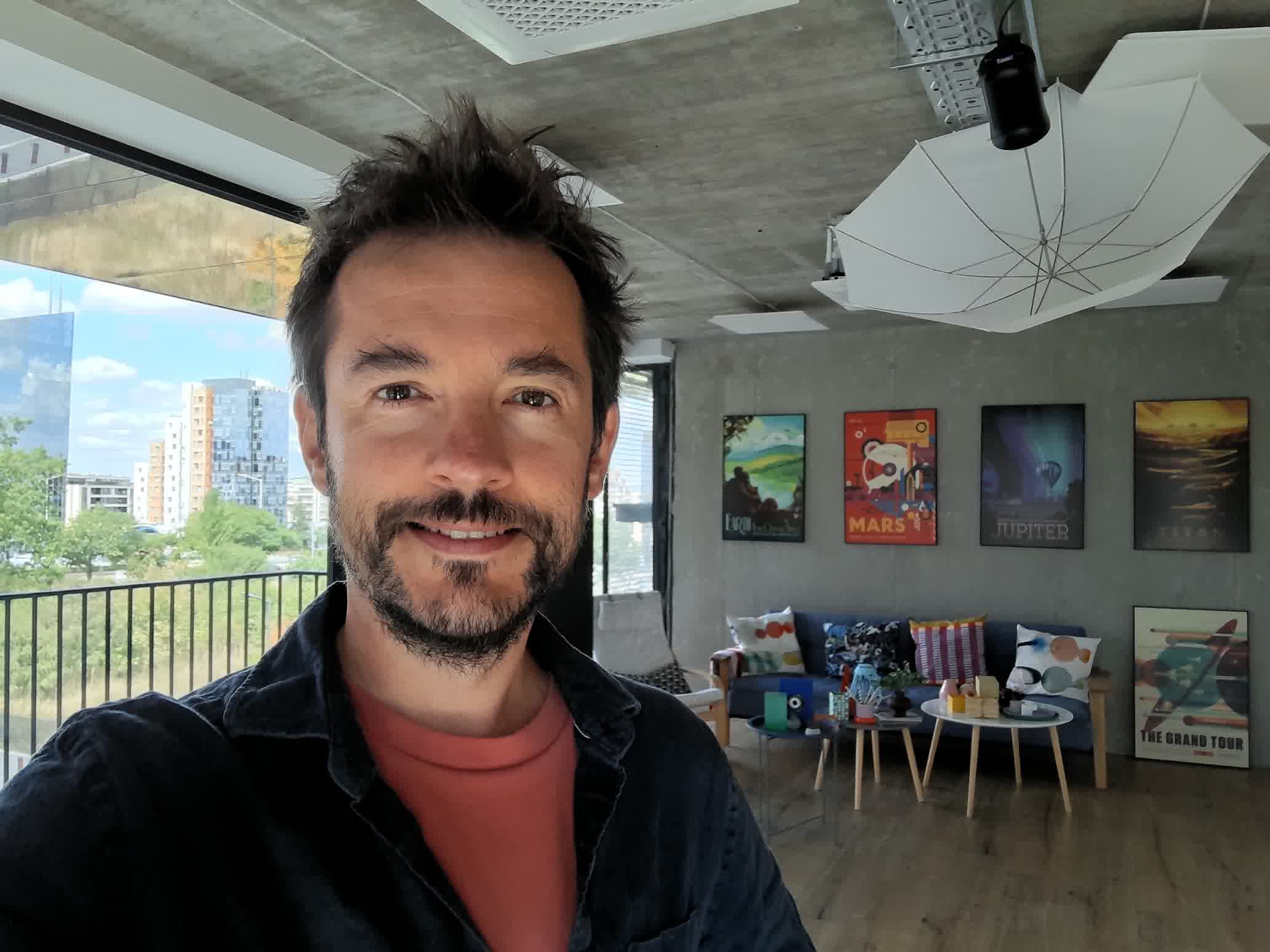 gsmarena_3001 Selfie of a smiling man with short dark hair and stubble indoors, with colorful posters on the wall behind him and a window on the left.