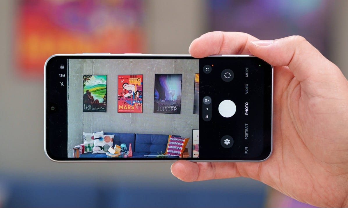 gsmarena_102 Hand holding a smartphone horizontally, filming a living room with colorful posters on the wall above a blue couch.