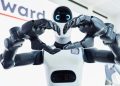 Close-up of two robotic hands forming a heart shape in front of a white robot head with blue glowing eyes.