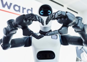 Close-up of two robotic hands forming a heart shape in front of a white robot head with blue glowing eyes.