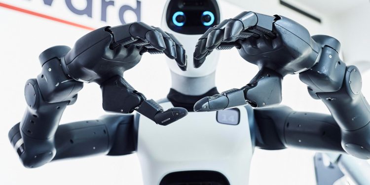 Close-up of two robotic hands forming a heart shape in front of a white robot head with blue glowing eyes.