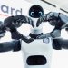 Close-up of two robotic hands forming a heart shape in front of a white robot head with blue glowing eyes.