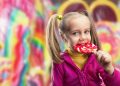 آیا باید در هزار روز اول زندگی کودکان را از قند دور نگه داریم؟ 17 Young girl in a pink jacket licking a red-and-yellow swirled lollipop in a bright candy shop background while smiling.