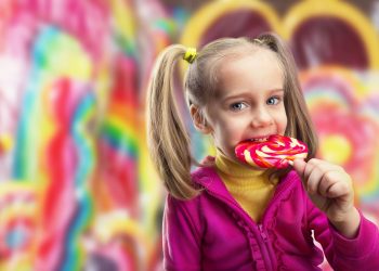 آیا باید در هزار روز اول زندگی کودکان را از قند دور نگه داریم؟ 13 Young girl in a pink jacket licking a red-and-yellow swirled lollipop in a bright candy shop background while smiling.