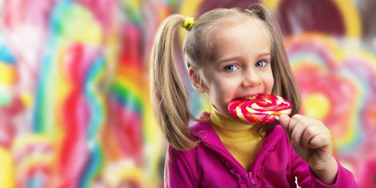 آیا باید در هزار روز اول زندگی کودکان را از قند دور نگه داریم؟ 1 Young girl in a pink jacket licking a red-and-yellow swirled lollipop in a bright candy shop background while smiling.