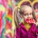 آیا باید در هزار روز اول زندگی کودکان را از قند دور نگه داریم؟ 8 Young girl in a pink jacket licking a red-and-yellow swirled lollipop in a bright candy shop background while smiling.