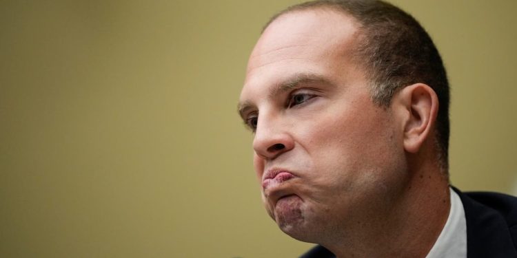 Close-up of a man in a suit making a tight-lipped, pouting expression (profile view).