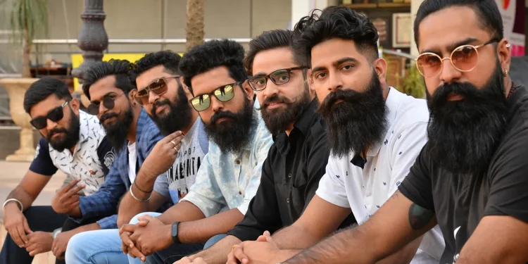 Seven men with beards and sunglasses sit in a row outdoors, posing for a group photo.
