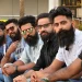 Seven men with beards and sunglasses sit in a row outdoors, posing for a group photo.