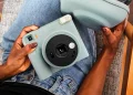 نمای بالای فردی نشسته با شلوار جین آبی که یک دوربین چاپ سریع آبی پاستلی Instax Square SQ2 و کیف محافظ هم‌رنگ آن را روی پای خود نگه داشته است.