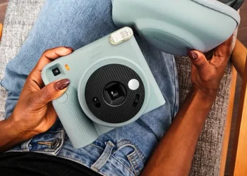 نمای بالای فردی نشسته با شلوار جین آبی که یک دوربین چاپ سریع آبی پاستلی Instax Square SQ2 و کیف محافظ هم‌رنگ آن را روی پای خود نگه داشته است.