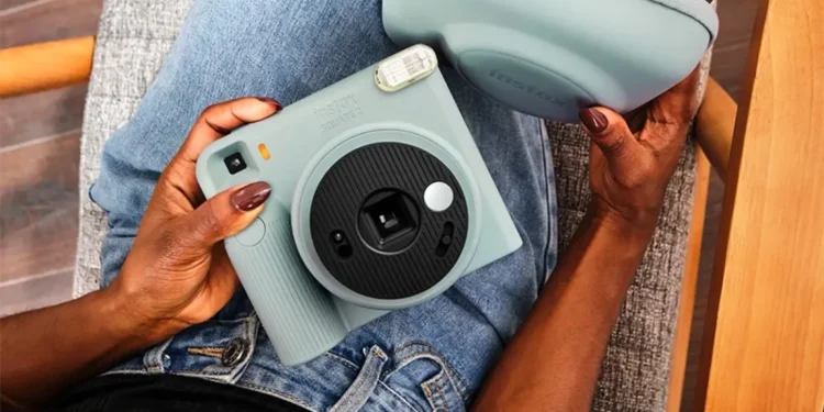 Fujifilm's Instax Square 2 camera was introduced with creative filters 1 نمای بالای فردی نشسته با شلوار جین آبی که یک دوربین چاپ سریع آبی پاستلی Instax Square SQ2 و کیف محافظ همرنگ آن را روی پای خود نگه داشته است.