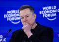 Man in a black jacket with hands folded near his chin on a World Economic Forum stage, speaking into a microphone.