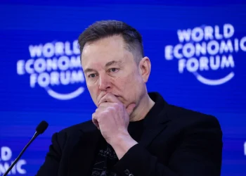 Man in a black jacket with hands folded near his chin on a World Economic Forum stage, speaking into a microphone.