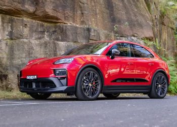 Red Porsche SUV (Macan) parked on a paved road beside a rocky wall with greenery in the background.
