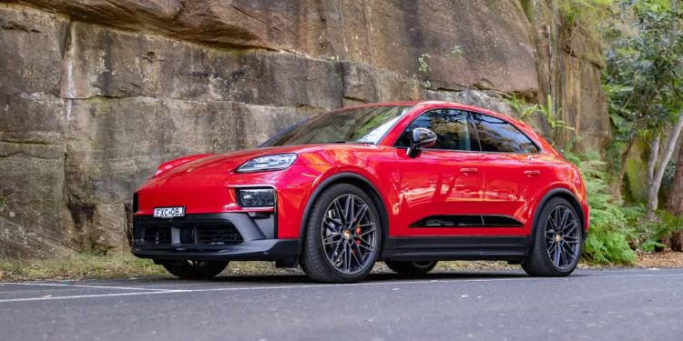 بررسی پورشه ماکان GTS 2026: خودرویی که شبیه سفینه فضایی است 1 Red Porsche SUV (Macan) parked on a paved road beside a rocky wall with greenery in the background.