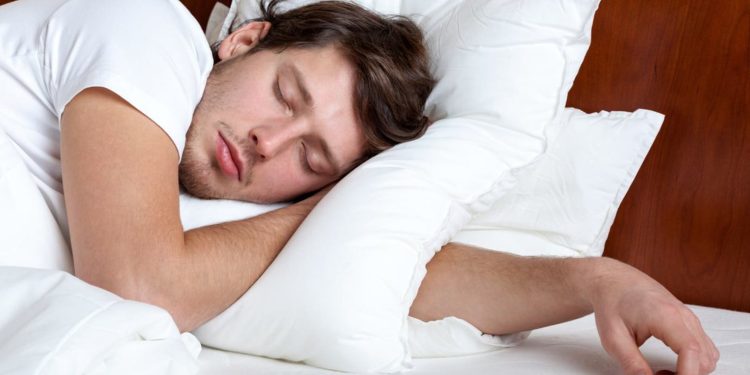 بهترین حالت خواب چیست؟ پاسخ سوال به شرایط بدن شما بستگی دارد 1 Young man sleeping on white pillows in a bed with a wooden headboard