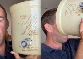 A man with short hair holds a large beige cylindrical storage tank with a side valve, shown in two frames side by side; he faces the camera in the left frame and drinks from the tank in the right frame (humorous prop).