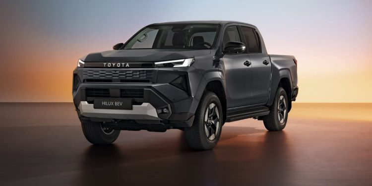 تماشا کنید: تویوتا هایلوکس برقی از سقوط یک کاروان 760 کیلوگرمی جان سالم به در می برد 1 Dark gray Toyota Hilux BEV pickup shown in a studio with a warm sunset gradient background, angled front-left view