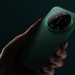 Hand holding a green smartphone with a large circular camera module on the back against a dark background, viewed from the side.