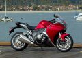 Red sport motorcycle parked on a wooden dock with a marina and boats in the background.