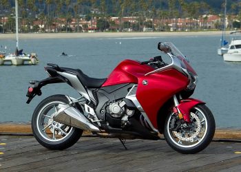 Red sport motorcycle parked on a wooden dock with a marina and boats in the background.