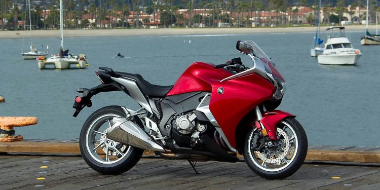 Red sport motorcycle parked on a wooden dock with a marina and boats in the background.