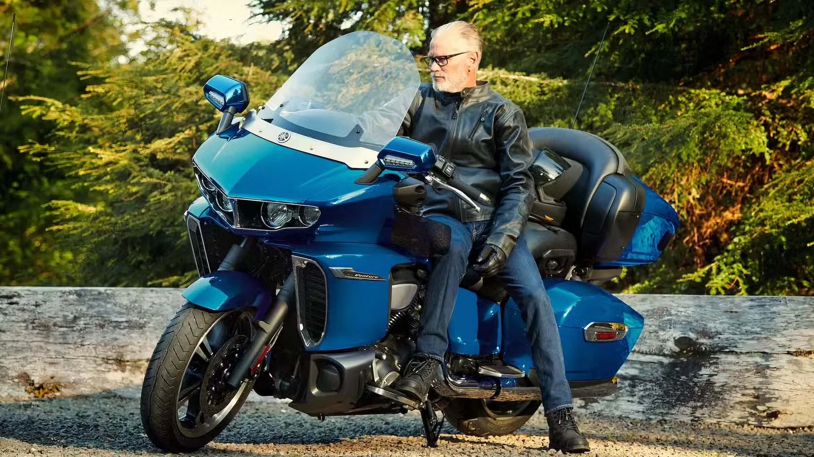 Man in a leather jacket sits on a large blue touring motorcycle with a windshield and storage, beside a wooden barrier.