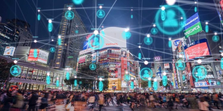 Nighttime city square with a crowded crosswalk, illuminated billboards, and teal floating user icons connected by white lines above the street, suggesting digital connectivity.