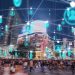 Nighttime city square with a crowded crosswalk, illuminated billboards, and teal floating user icons connected by white lines above the street, suggesting digital connectivity.